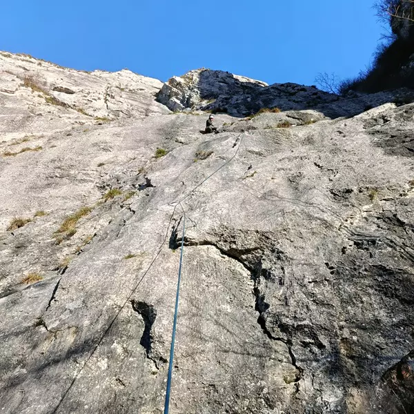 Een bergbeklimmer beklimt een steile, rotsachtige rotswand onder een helderblauwe hemel, terwijl een touw langs de rots naar beneden loopt, in de richting van de camera.